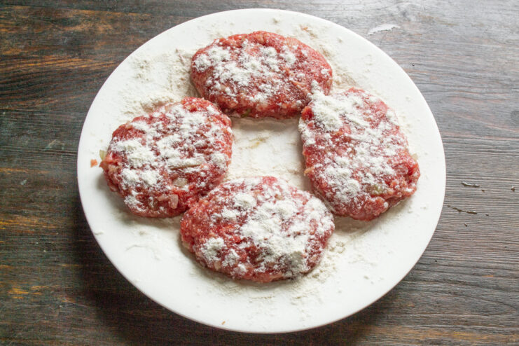 Forming the Patties and Coating in Flour Four round, uncooked ground beef patties coated in flour on a wooden board.