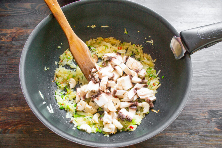 Adding Mushrooms to the Sautéed Onions Diced mushrooms being added to a pan with sautéed onions.