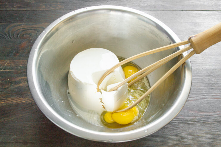 Preparing the base for the zapekanka: ricotta, eggs, and salt in a mixing bowl. A white bowl containing ricotta cheese, two cracked eggs, and a pinch of salt, ready for mixing.