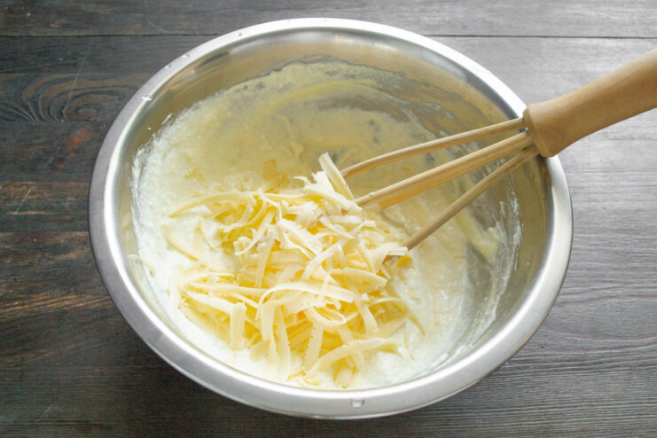 Adding grated cheese to the ricotta and egg mixture for a rich flavor. Hand adding a portion of grated yellow cheese to the ricotta and egg mixture in a white bowl.
