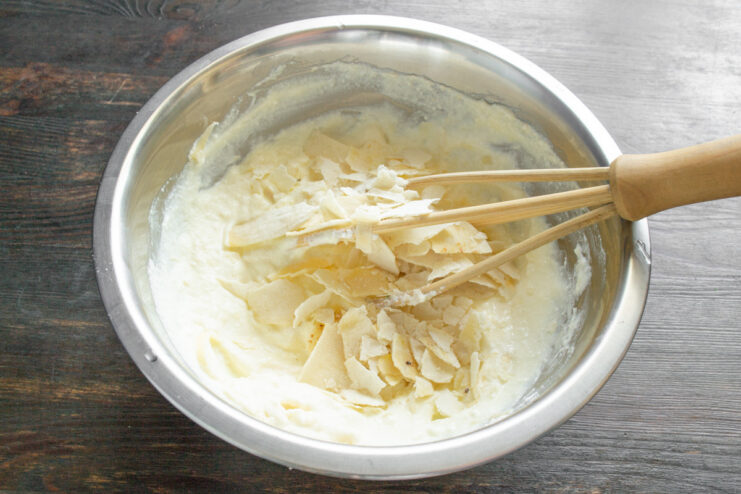 Whisking the ingredients together to create a smooth zapekanka batter. A whisk mixing the cheese, ricotta, and egg batter in a white bowl.