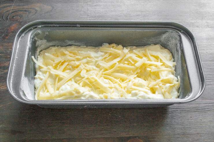 Assembling the zapekanka in a loaf pan, topped with cheese and ready for the oven. Zapekanka batter in a loaf pan, topped with a generous layer of grated cheese before baking.