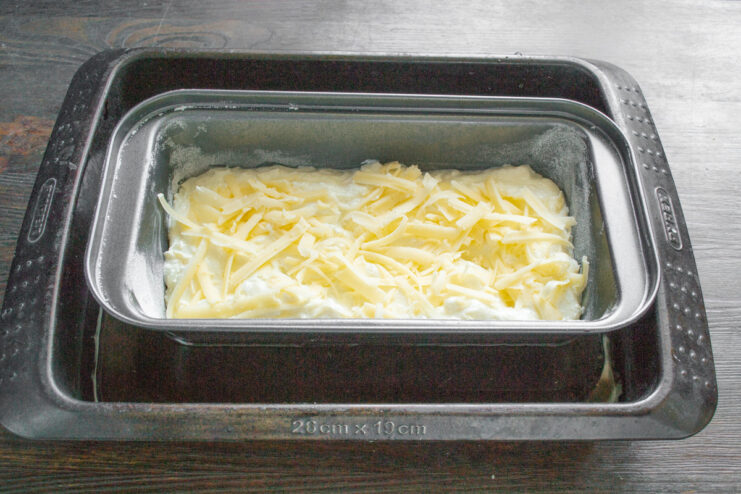 How to set up a water bath (bain-marie) for a perfectly moist zapekanka. A loaf pan with zapekanka batter placed inside a larger baking dish, ready for a water bath.