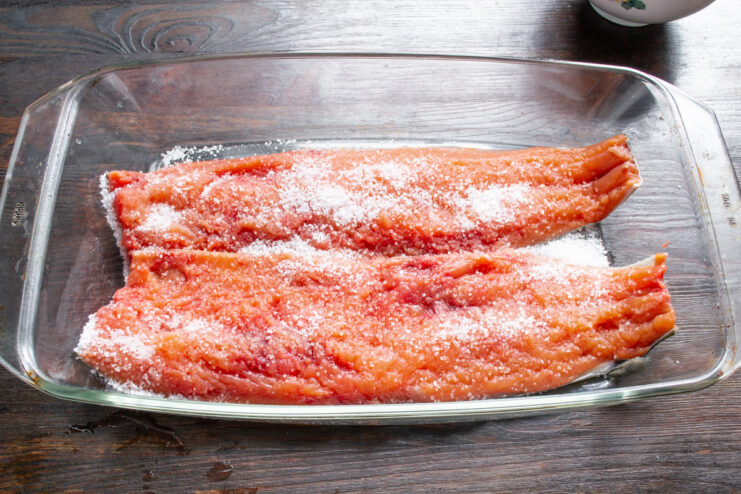 Applying the Salt and Sugar Cure to the Salmon Hand sprinkling a mixture of salt and sugar over a pink salmon fillet in a glass dish.