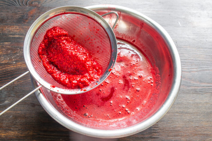 How to make smooth raspberry puree for zefir Straining raspberry puree through a fine-mesh sieve into a white bowl.