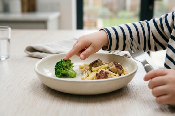 A child's hand tentatively touching a broccoli floret resting on a plate next to pasta