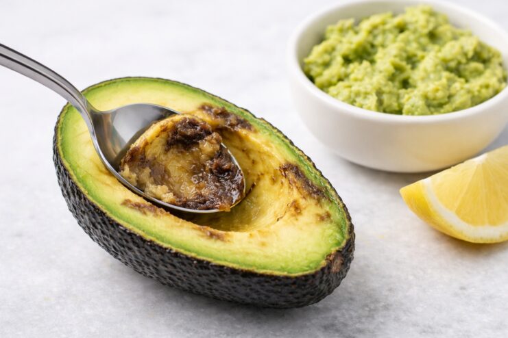 Scooping Out Overripe Avocado Flesh Metal spoon scooping out dark brown spots from a halved avocado next to a fresh lemon