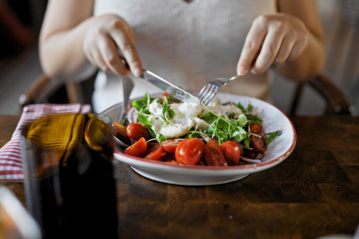 A crosswise cut on a fresh burrata cheese to reveal the creamy stracciatella interior. A hand gently slicing a crosswise opening in a fresh burrata cheese on a salad plate.