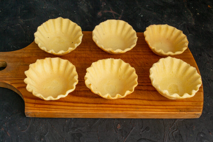Cooling the Tartlet Shells Six empty shortbread tartlet shells on a cutting board