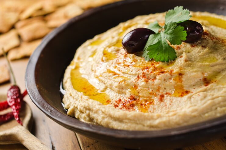 Silky Lebanese Style Hummus Ultra smooth hummus topped with olives and fresh parsley presented in a dark ceramic bowl.