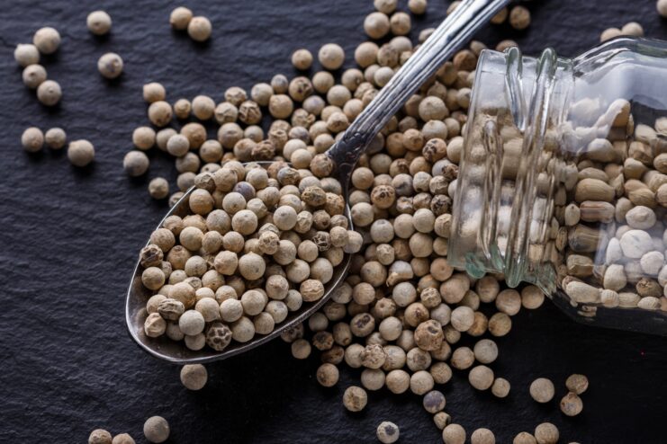 Whole white peppercorns scattered from a small glass jar onto a dark surface.
