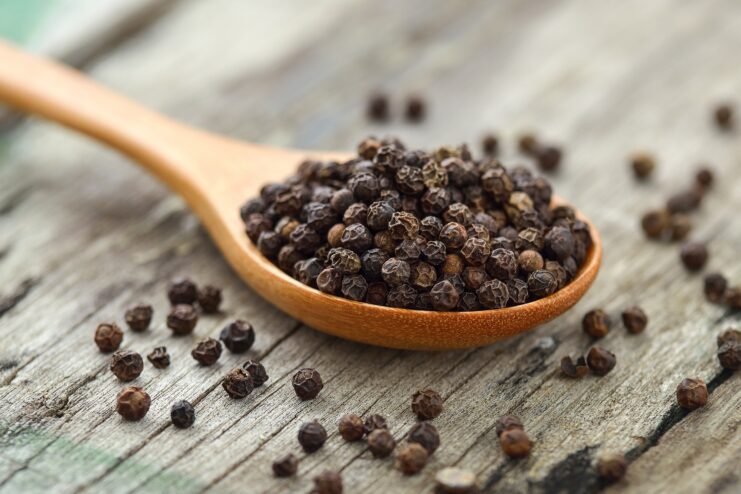 Close-up view of whole black peppercorns resting in a wooden spoon on a wooden table.