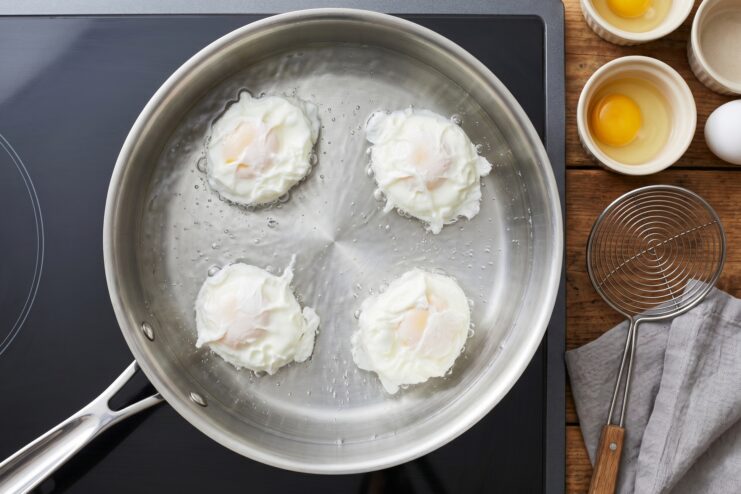 Poaching Multiple Eggs Four perfectly shaped poached eggs simmering together in a wide stainless steel pan filled with calm, hot water.