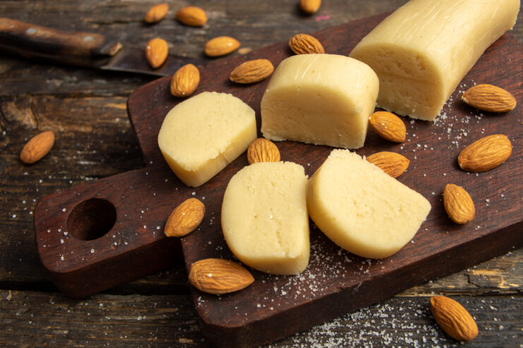 Homemade elastic marzipan log resting on a wooden board surrounded by whole almond kernels