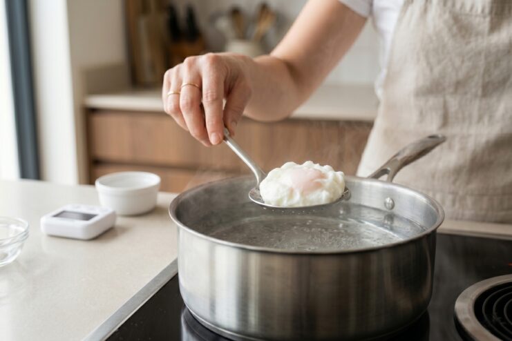 Checking Poached Egg Doneness Cooked poached egg resting on a stainless steel slotted spoon held over a pot of hot water.
