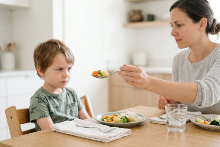 Parent Forcing a Child to Eat Mother attempting to spoon-feed vegetables to a visibly displeased little boy sitting at the dinner table.