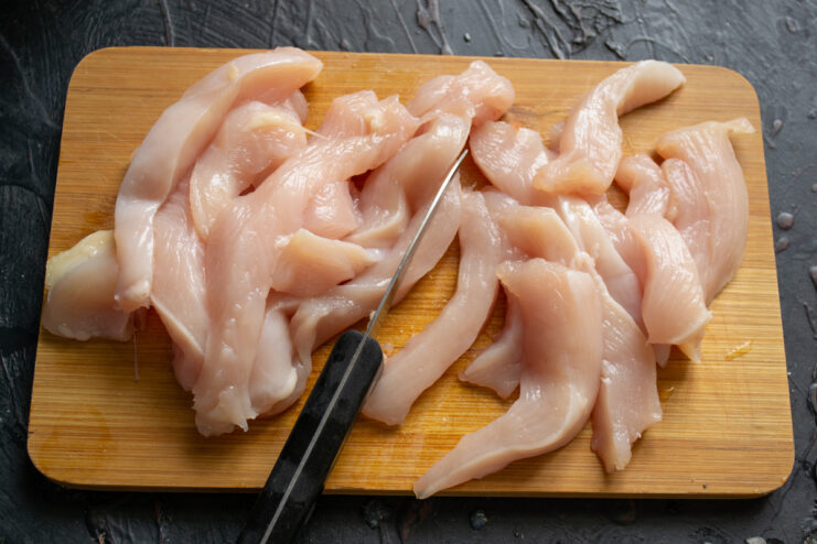 Prepping The Chicken Slicing raw chicken breast into thin strips on a cutting board