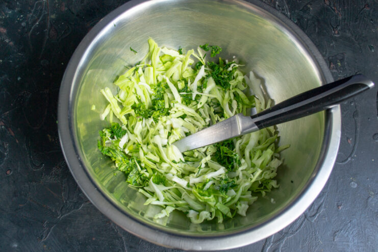 Preparing the Cabbage Base Freshly shredded green cabbage mixed with chopped dill in a metal mixing bowl.