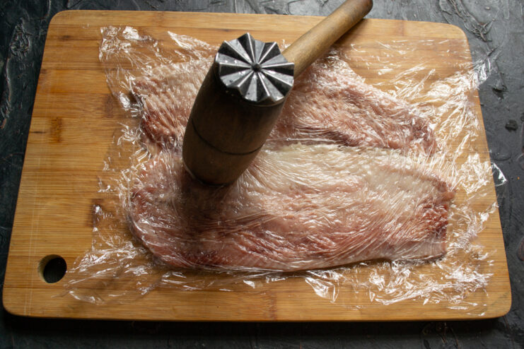 Pounding herring fillets with a meat mallet through plastic wrap on a wooden board.