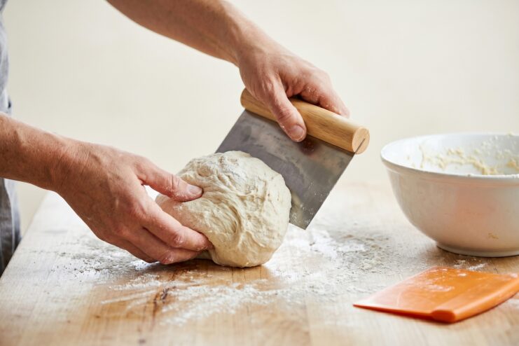 Metal bench scraper cutting dough Chef using a professional metal bench scraper to fold and knead sticky dough on a wooden counter.