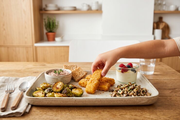 Proper Presentation of Healthy Meals for Children Child taking a fish stick from a plate alongside roasted Brussels sprouts, mushrooms, and a small serving of pâté.