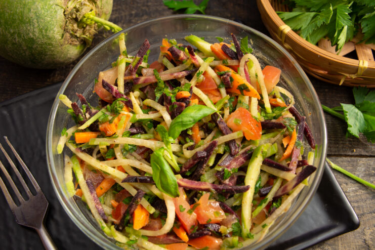 Green radish salad with purple carrots and tomatoes in a glass bowl
