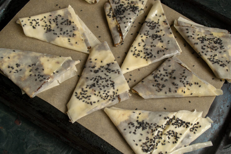 Adding Sesame Seeds Samosas on a baking sheet sprinkled with black sesame seeds before baking