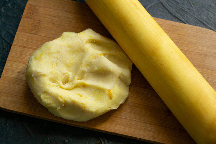 A ball of shortcrust pastry on a wooden board next to a yellow rolling pin.