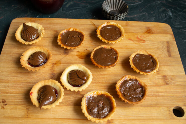 Piping the chocolate buttercream over the jam Chocolate buttercream piped over the jam into the shortcrust pastry shells