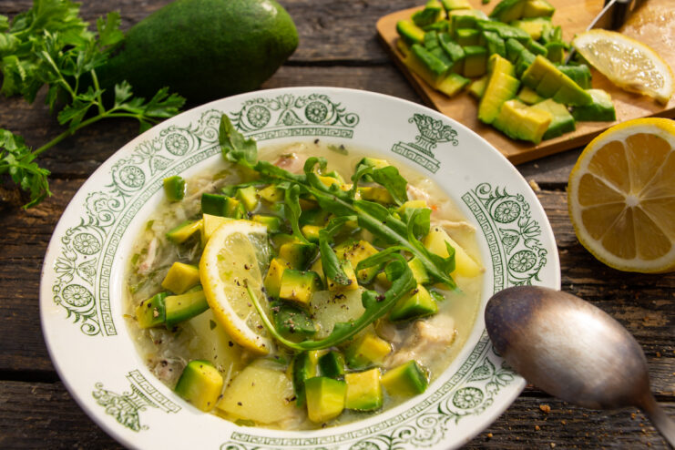 Finished chicken soup with diced avocado, a lemon slice, and fresh arugula served in a bowl