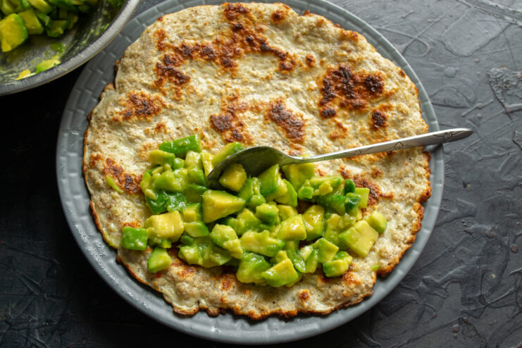 Assembling The Healthy Breakfast Spoonfuls of dressed avocado being placed onto one half of a cooked oat pancake