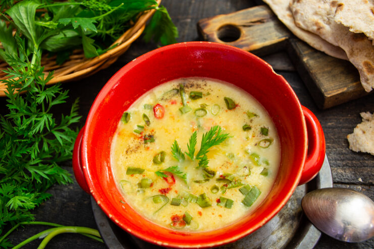 Appetizing cheese and vegetable soup in a red bowl garnished with herbs and served with flatbread
