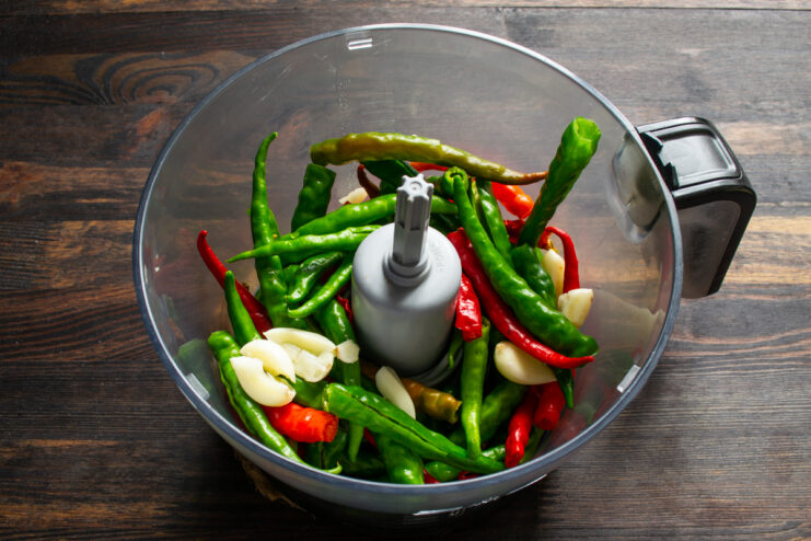 Ingredients in a food processor Whole pepper pods and garlic cloves in the clear bowl of a food processor.