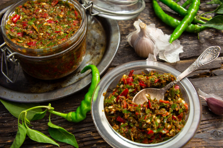 Ready raw adjika in a glass jar and a bowl with chili peppers and garlic in the background.