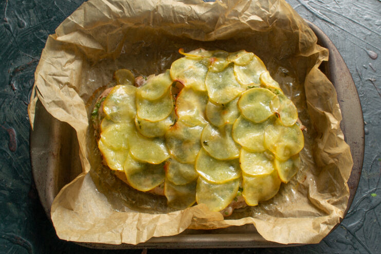Golden baked chicken covered in potato slices on a baking sheet