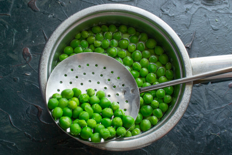 Scooping boiled green peas from a pot with a slotted spoon