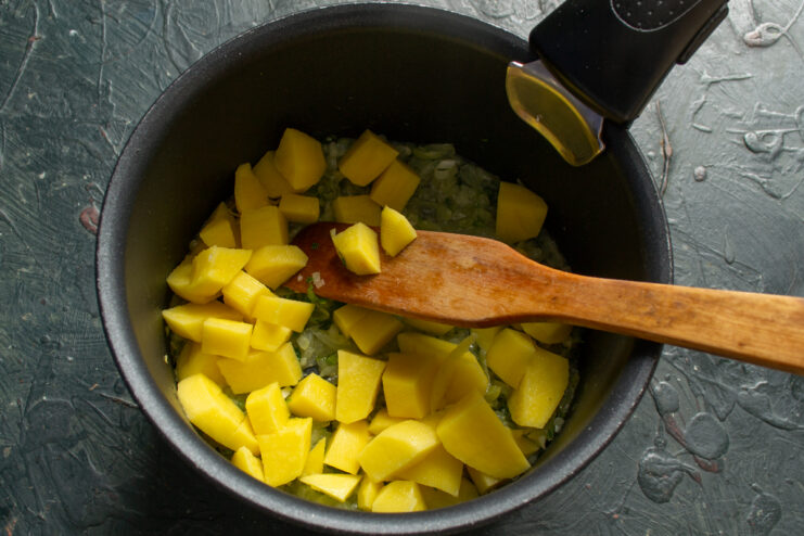 Small raw potato cubes being added to the sauteed vegetable mix in a cooking pot.