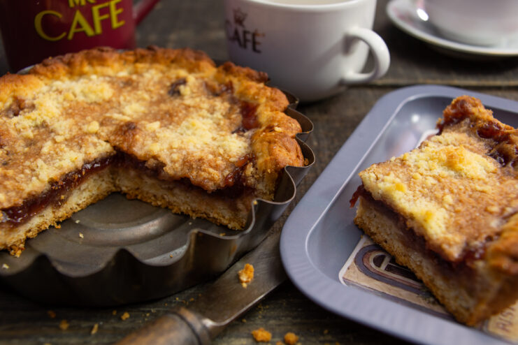 Slice of strawberry jam pie with streusel topping on a plate next to a cup of coffee.