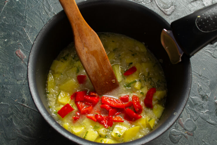 Chopped pieces of sweet red bell pepper being stirred into a pot of cooking potatoes.