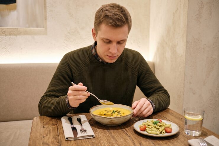 Consuming simple and convenient food during anxiety A man eats soup with a spoon at a cafe table, with a plate of penne pasta nearby.