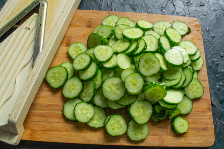 Slicing the Cucumbers Thinly sliced cucumbers on a cutting board next to a mandoline slicer