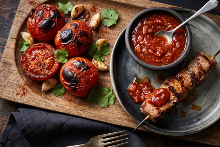 Char-roasted tomatoes and garlic placed next to a bowl of thick tomato sauce and grilled meat skewers.