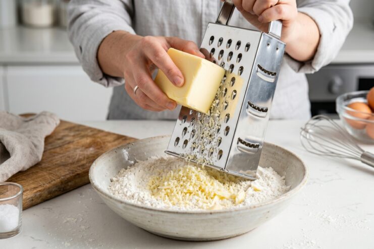 Grating Frozen Butter for Dough A chef's hands grating a block of frozen butter on a coarse grater into a bowl of flour.