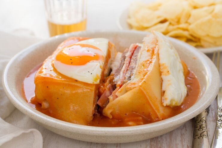 Layered Francesinha sandwich covered in melted cheese, topped with an egg, and surrounded by sauce