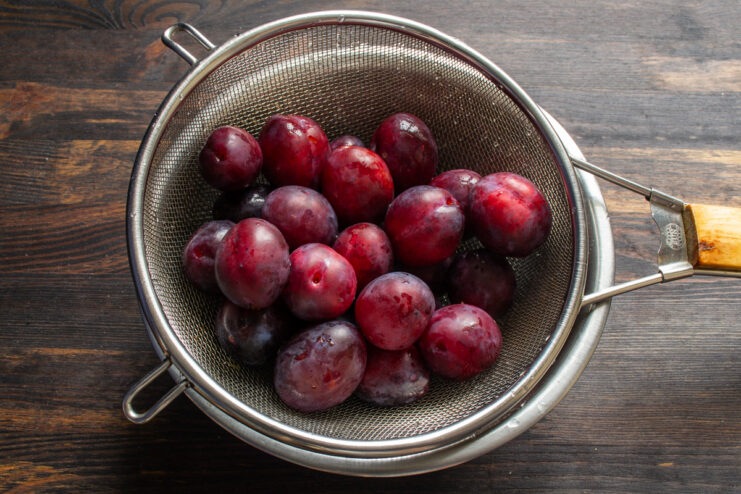 Washed ripe red plums resting inside a metal wire strainer