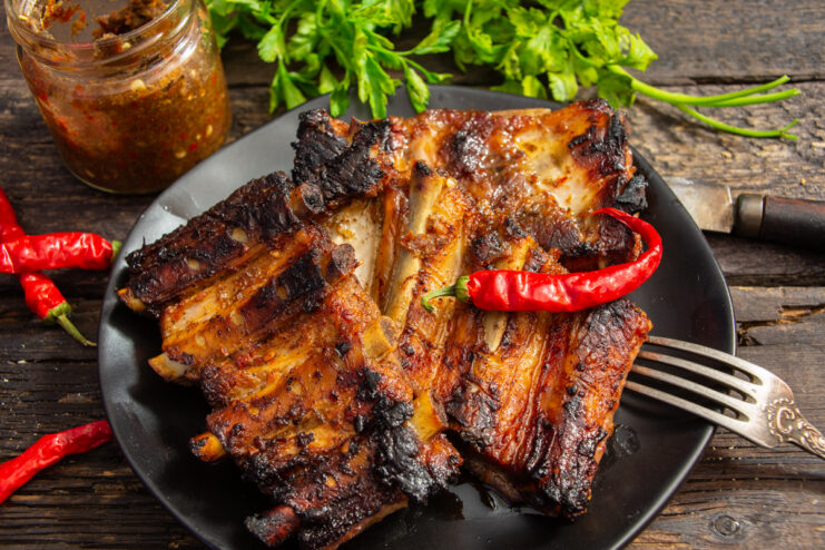 Baked pork ribs with adjika garnished with fresh herbs and hot peppers on a plate.