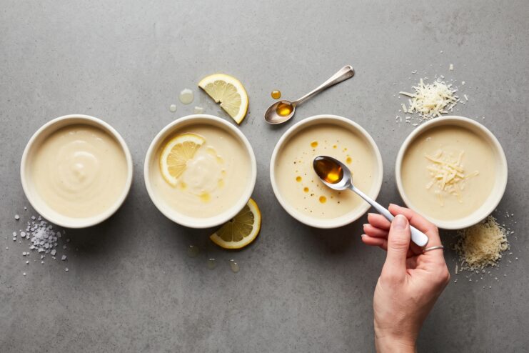 Correcting and balancing dish flavors Top view of four bowls of cream soup being seasoned with lemon juice, oil, grated cheese, and salt to balance flavors.
