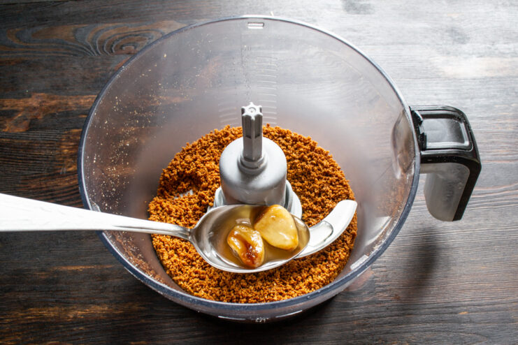 Adding garlic and olive oil Adding roasted garlic cloves to crushed peanuts in a blender