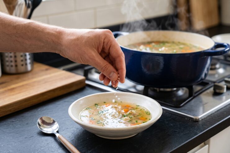Adding salt to simmering soup Cook dropping a pinch of coarse salt into a bowl of soup with a simmering pot in the background.