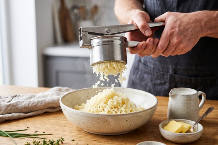 Pressing boiled potatoes through a stainless steel ricer into a deep mixing bowl.